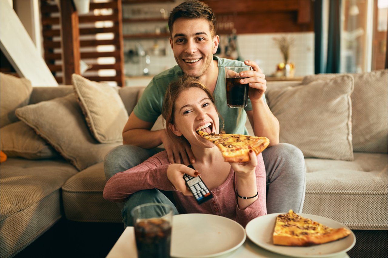 Pärchen ist Pizza vor dem TV und achtet nicht auf die Mahlzeit. So bekommt man schneller wieder Hunger und nimmt nicht ab.